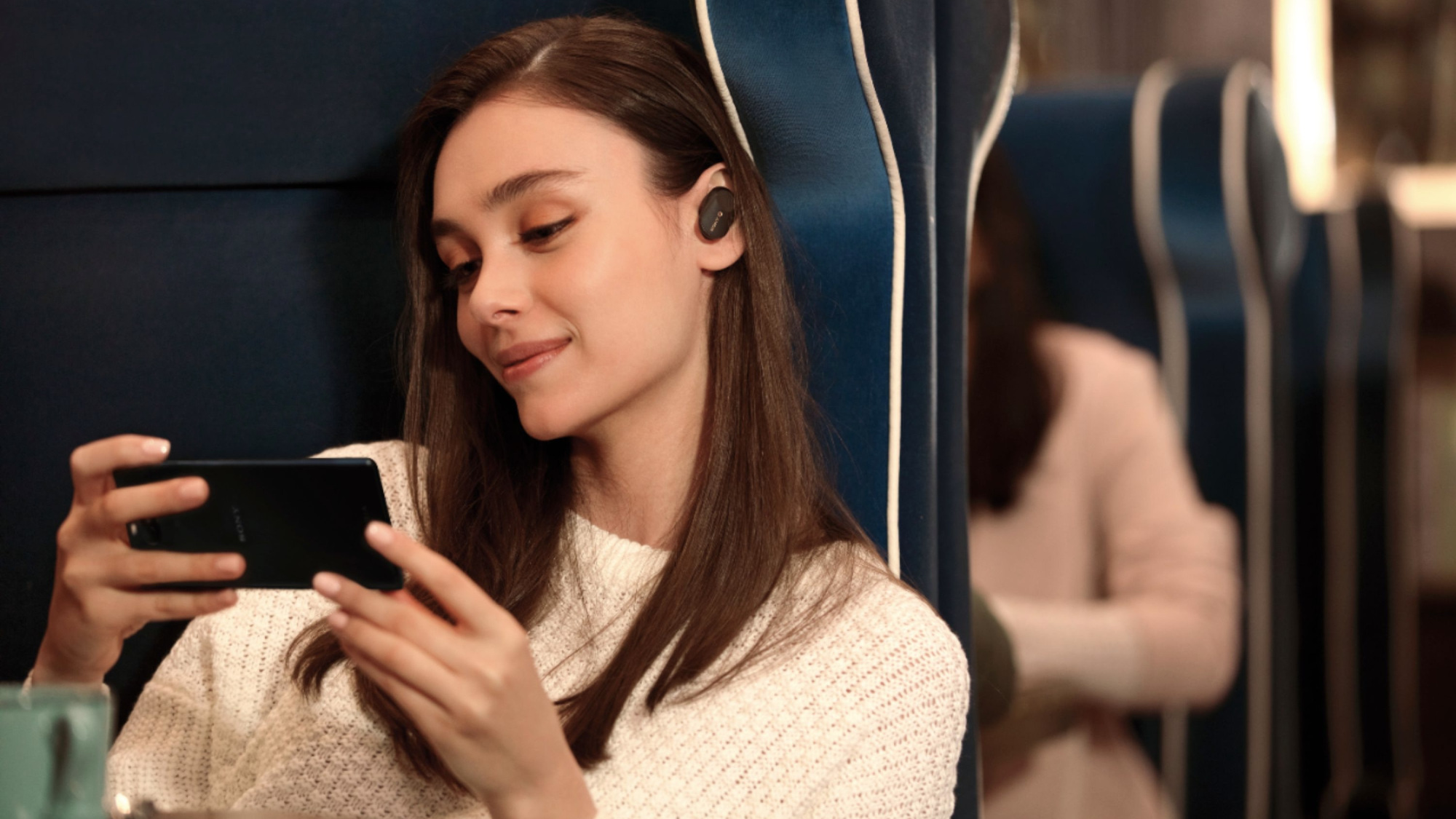 a woman sitting on a train watches a video on her phone while listening with her earbuds