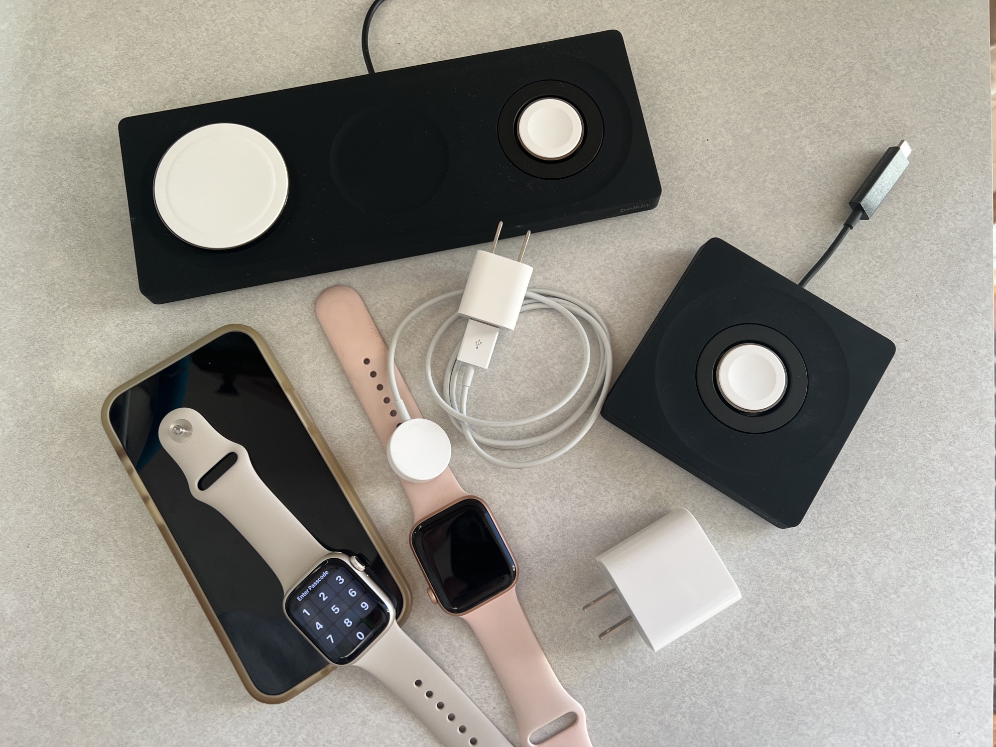 A collection of chargers, including the Belkin 3-in-1 pad, the Belkin Apple Watch Charger, the Apple Watch charger, a 20 watt USB-C adaptor, an iPhone 12, and two Apple Watches.