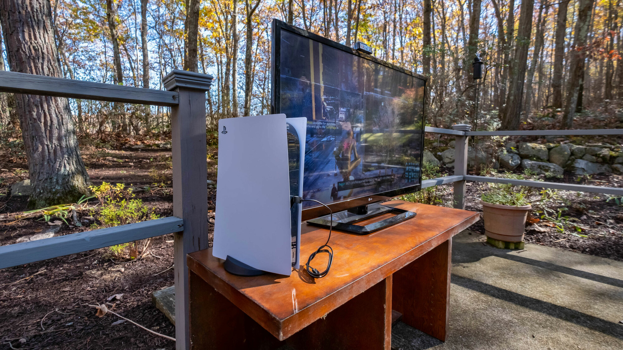 A PlayStation 5 sitting next to a TV, both set up on an entertainment center that's outdoors with a wooded setting as the backgdrop.