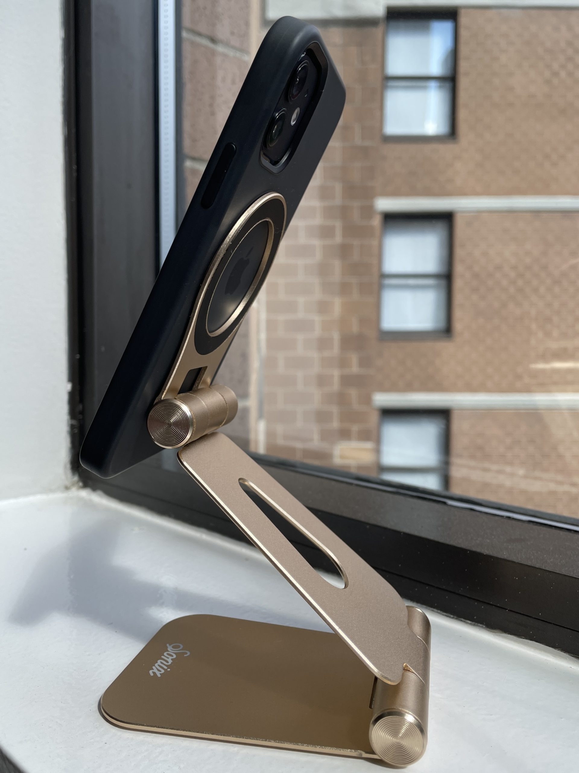An iPhone 12 attached via magnets to a gold finish metallic phone stanc, shaped like an S.