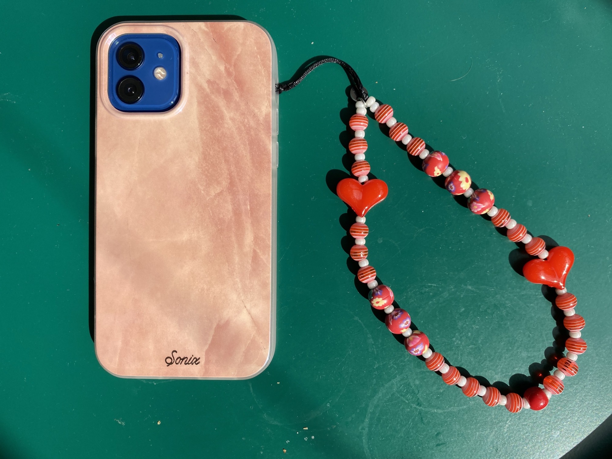 An iPhone 12 in a pink case attached to a pink beaded phone charm via the volume button hole in the case.