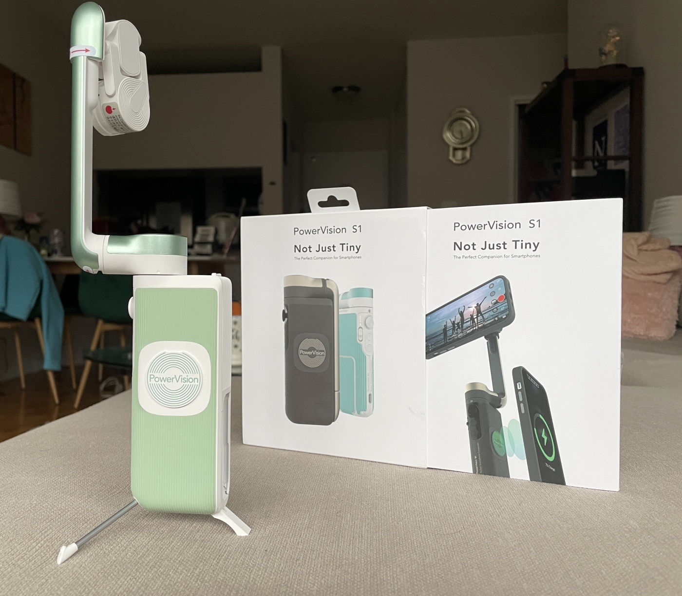 A small green camera gimbal, with three small legs set up for a tripod on the bottom, next to its product box. 