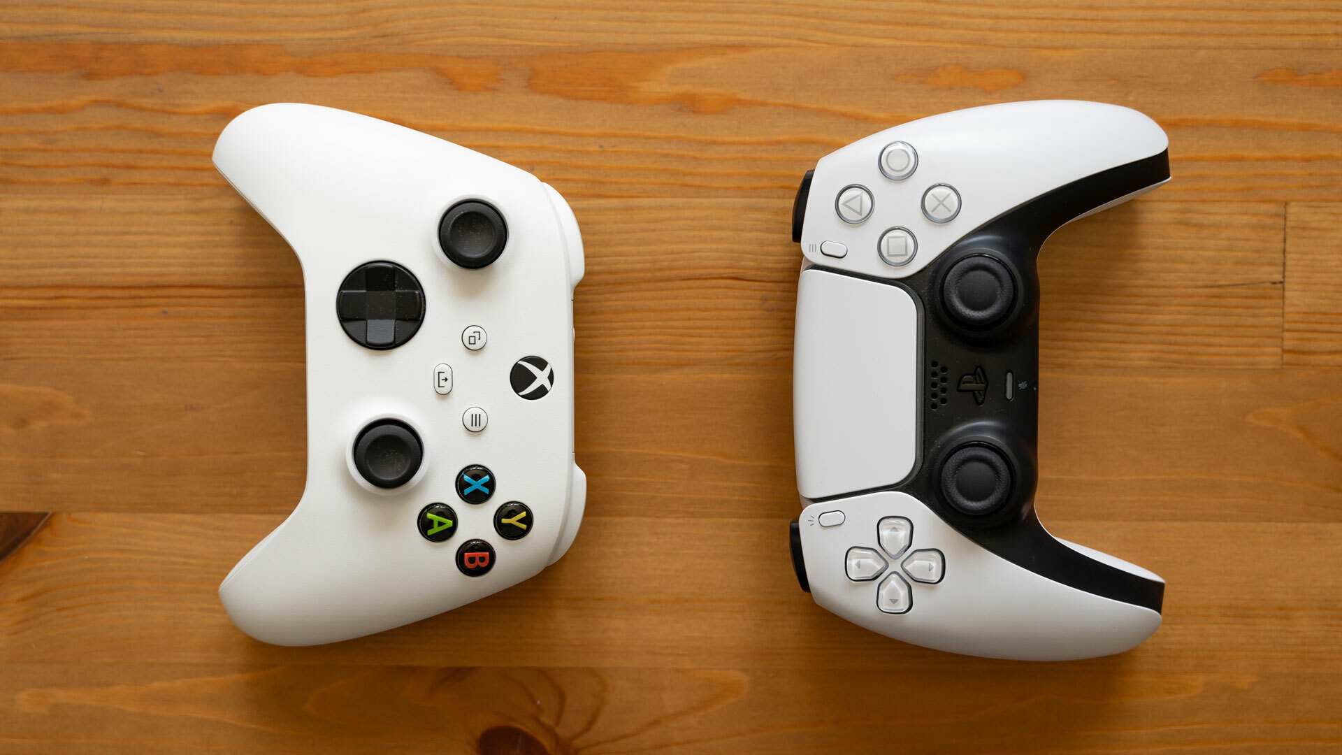 A white Xbox Series controller an a PlayStation 5 DualSense controller sitting side by side on a wooden surface.