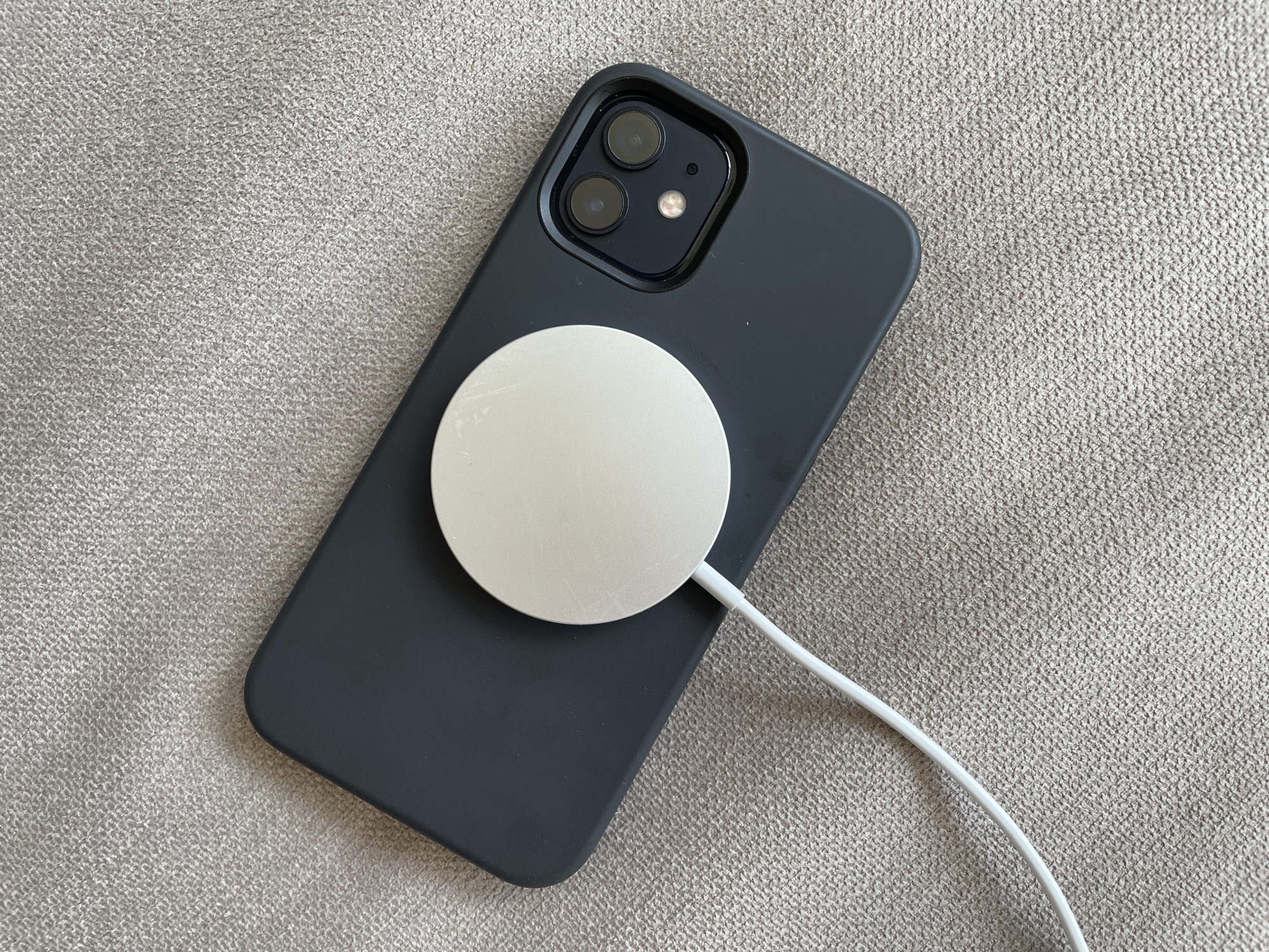 An iPhone 12 in a black case attached to Apple's MagSafe wireless charger, a round aluminum puck stuck on via magnets.
