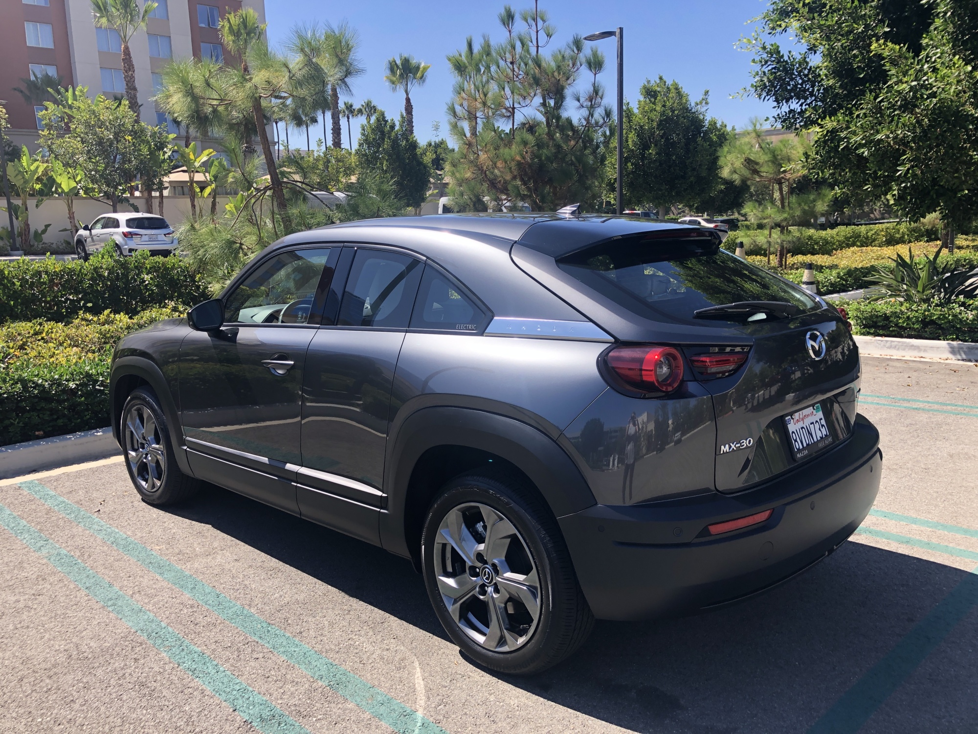 A silver car in a parking lot with green trees and bushes behind it. 