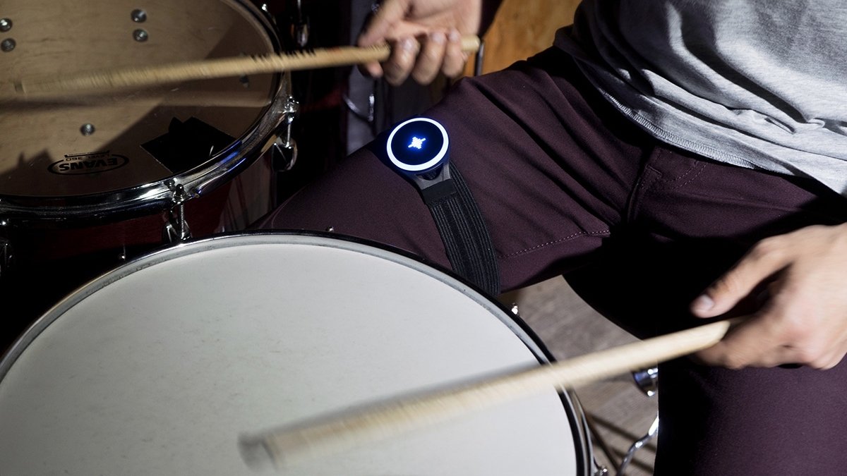 Two hands holding drumsticks above snare drum with legs on either side of it, one of which has blue glowing gadget strapped to thigh