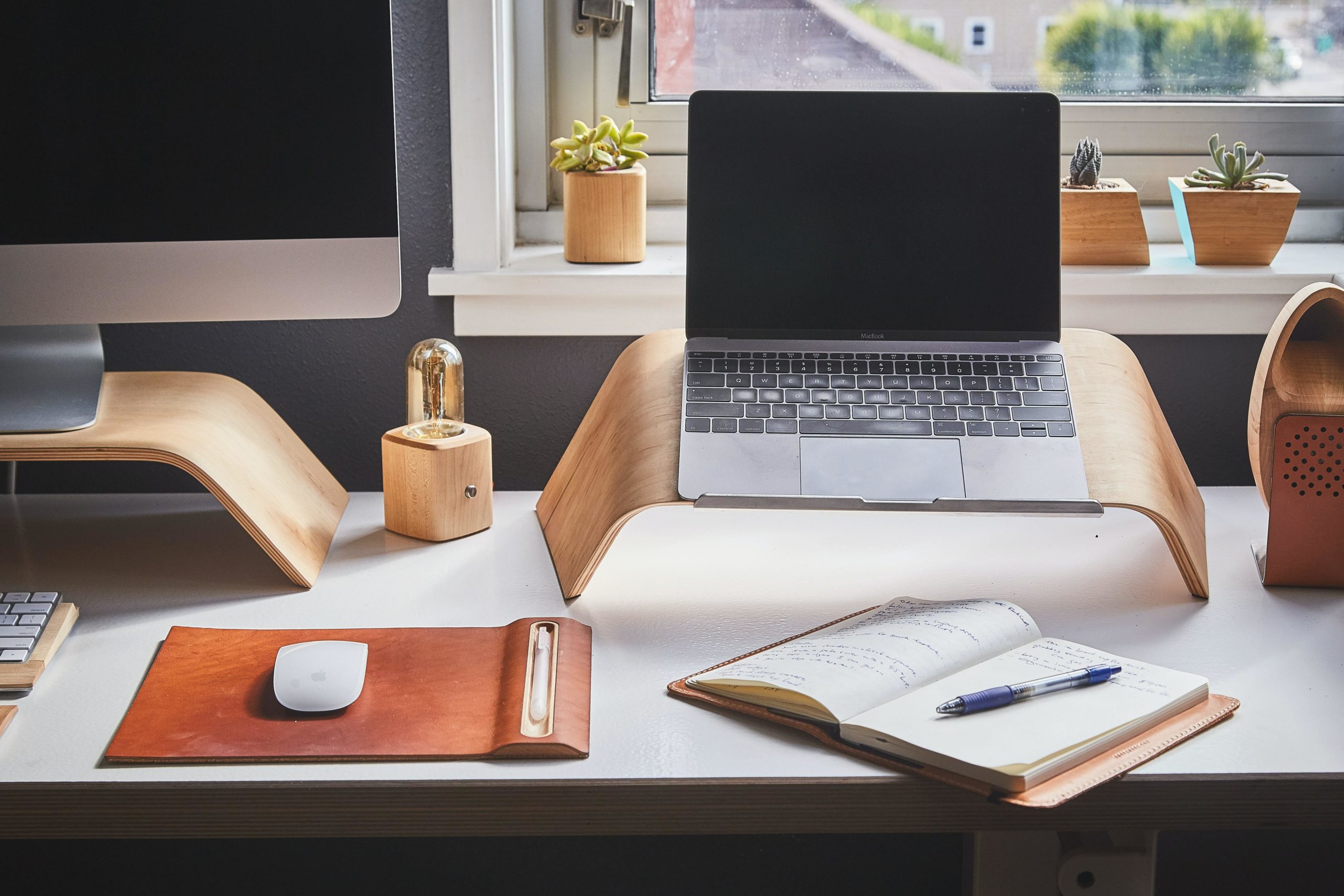 A laptop sits atop a stand on a desk next to a sunny window. A notepad and pen sit beside a mouse on a mousepad.