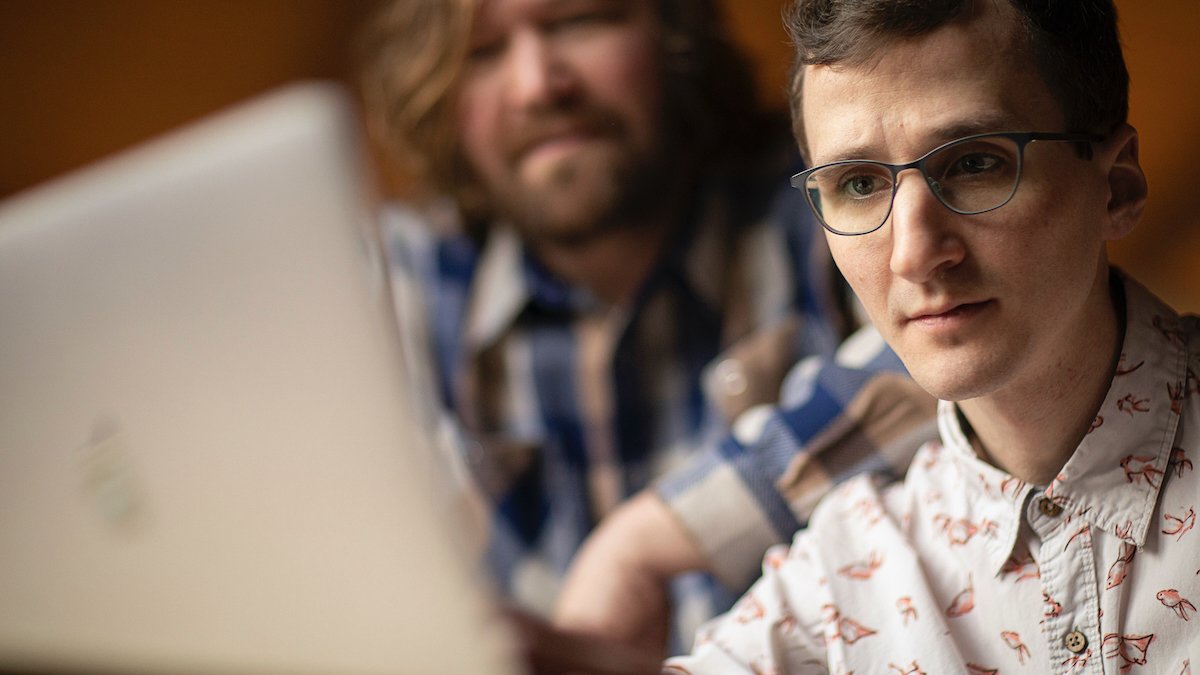 Person wearing glasses looking at Macbook with other person out of focus behind him