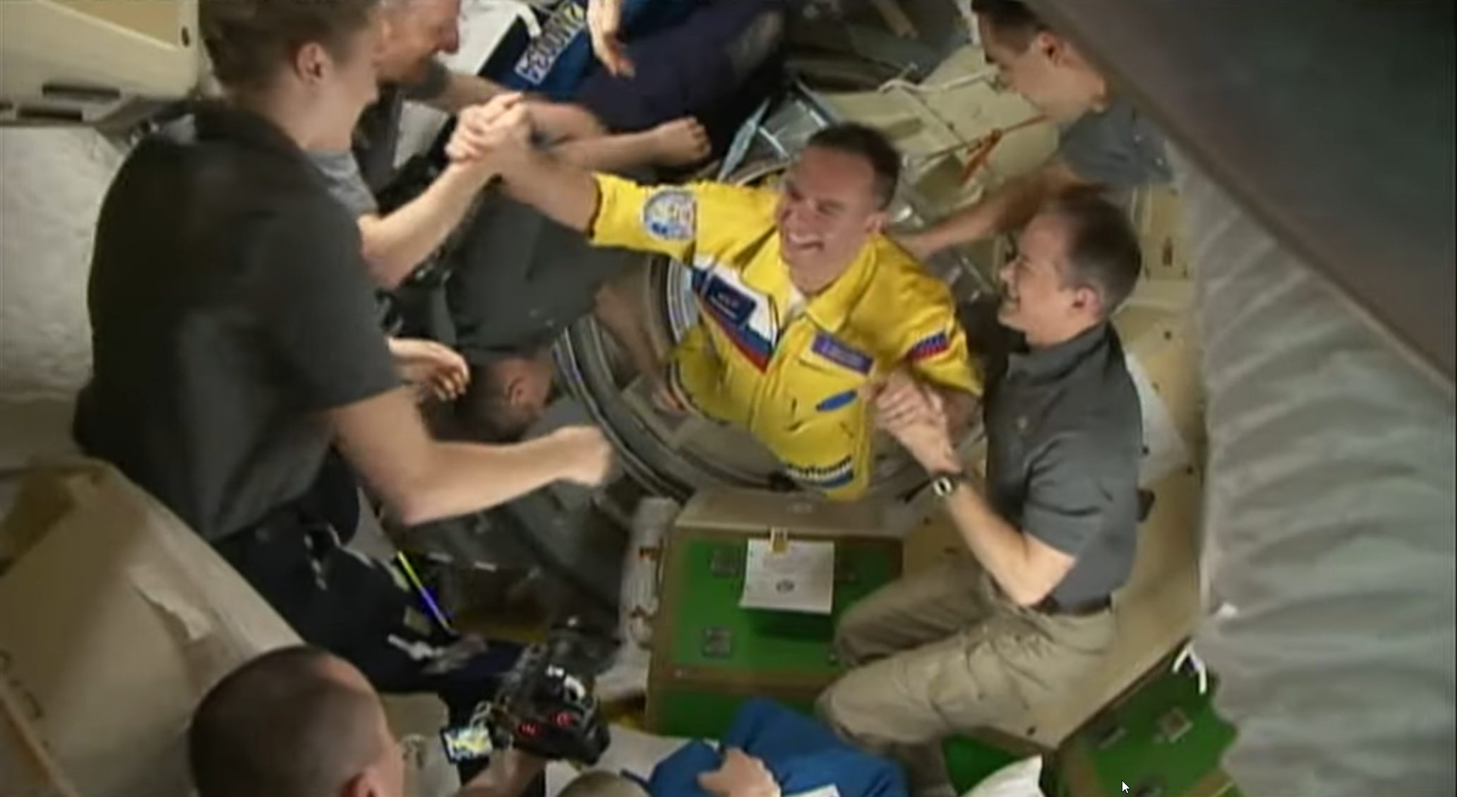A smiling cosmonaut wearing a yellow flight suit with blue accents floats out of an open hatch as astronauts aboard the International Space Station greet him.