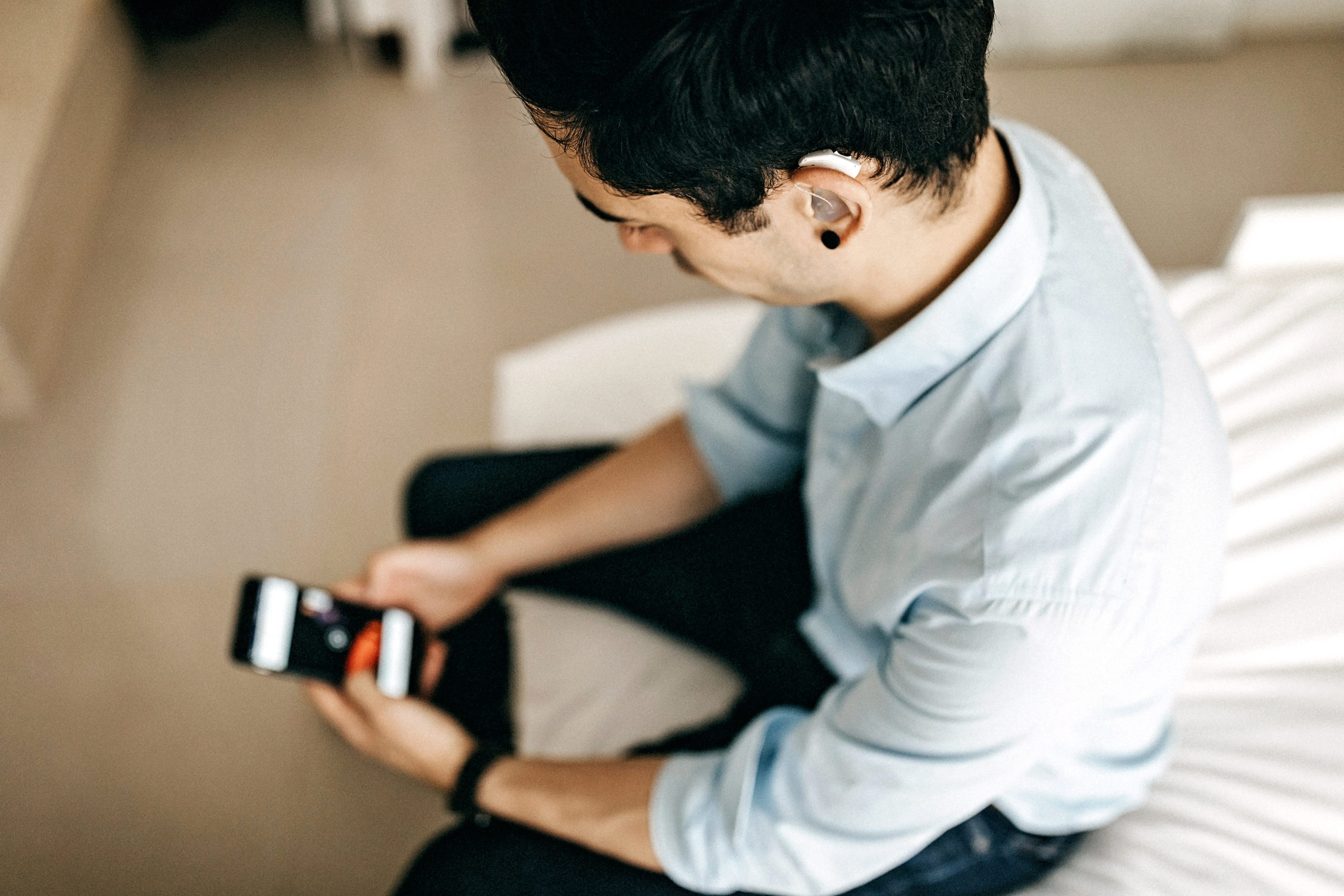 A person with a hearing aid sits on a bed using their cell phone.