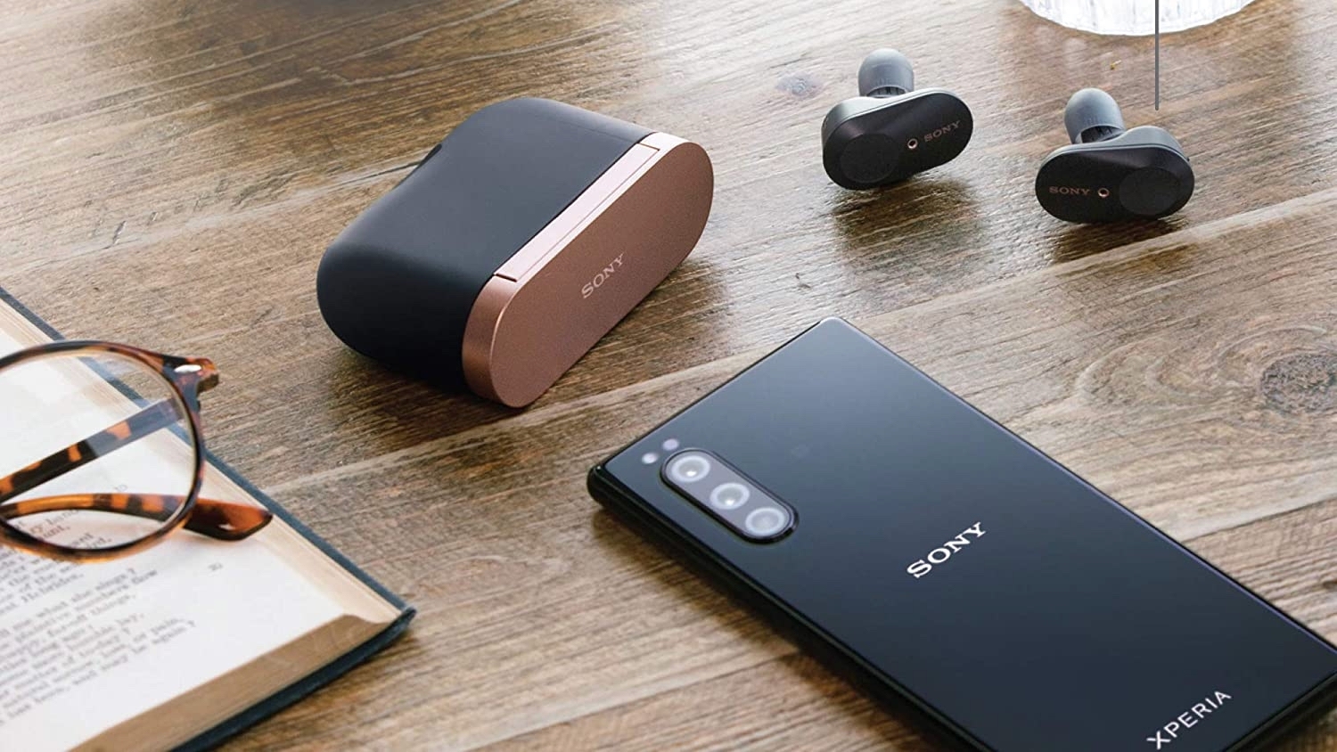 Black earbuds with black and rose gold case on wooden table with black sony phone, book and glasses,  and glass of carbonated drink 