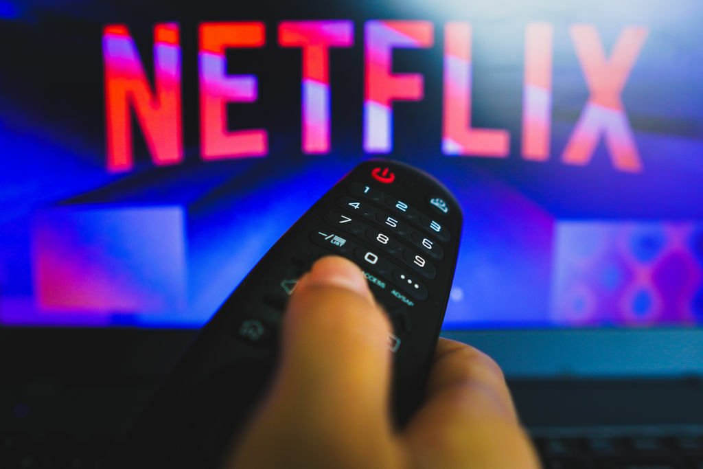 A hand holding a remote control, pointing at a screen with the Netflix logo.