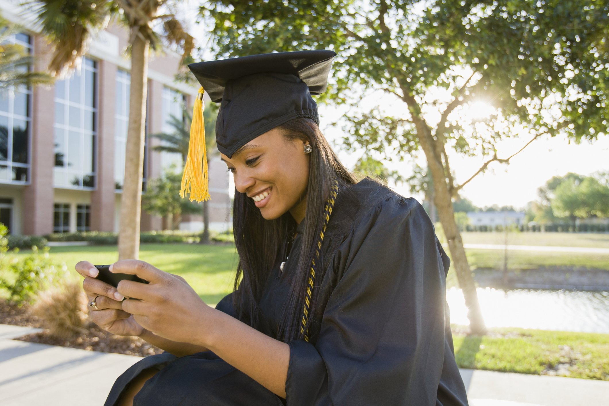 Young woman in graduation cap and gown sits on ground on her phone