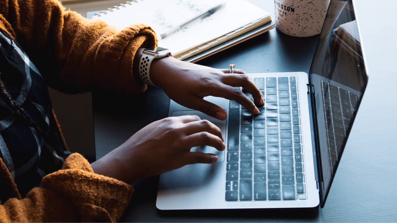 a woman typing on a laptop that's next to a notebook and a cup of coffee. she's wearing a yellow sweater, a smartwatch, and a diamond ring