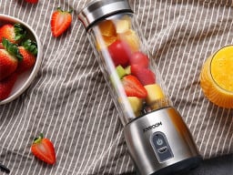 Silver juicer with cut-up fruits inside it next to glass of juice and bowl of strawberries