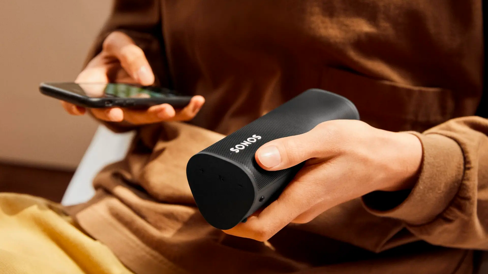 a close-up of a person in a brown outfit holding a sonos roam speaker and a smartphone