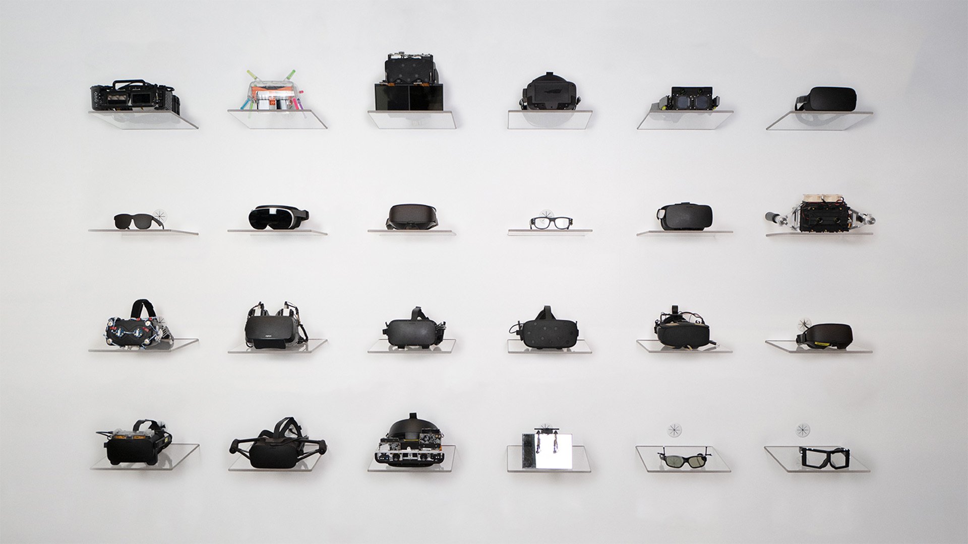 24 prototypes of varying sophistication sit on a white wall at Reality Labs.