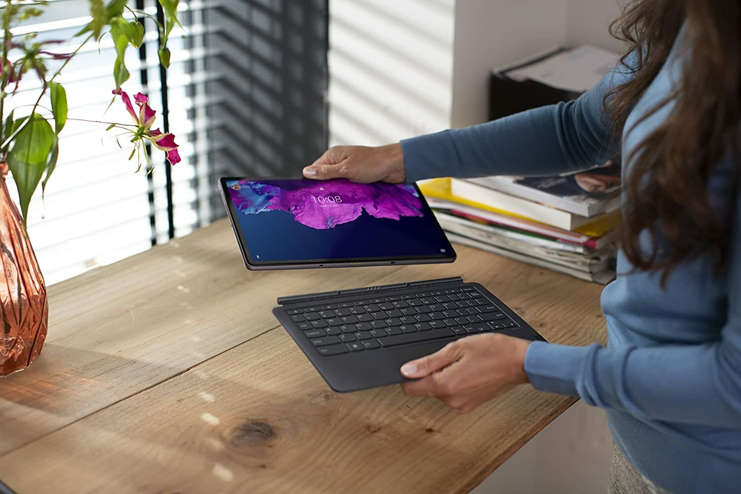 Woman standing next to desk holding Lenovo Tab P11 Plus tablet with a detachable keyboard.