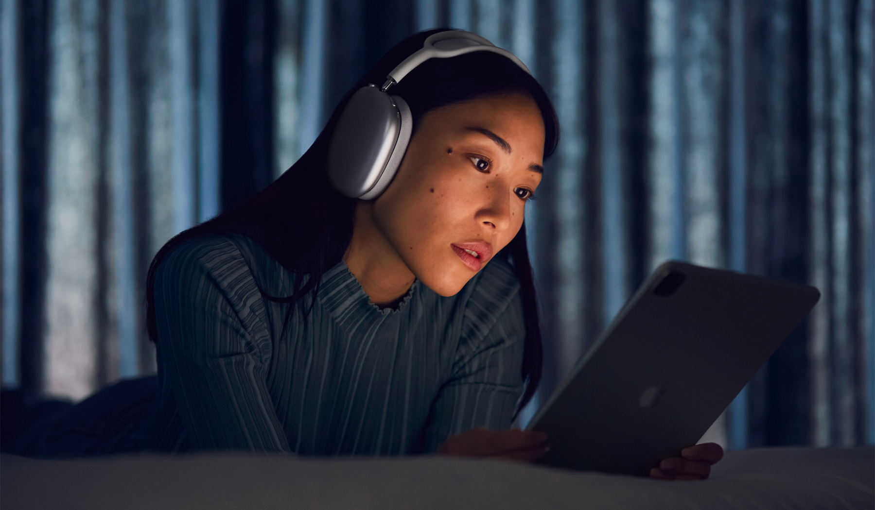 a woman in a striped turtleneck laying on a bed in a dark room. she's wearing a pair of blue airpods max and looking at a lit-up ipad
