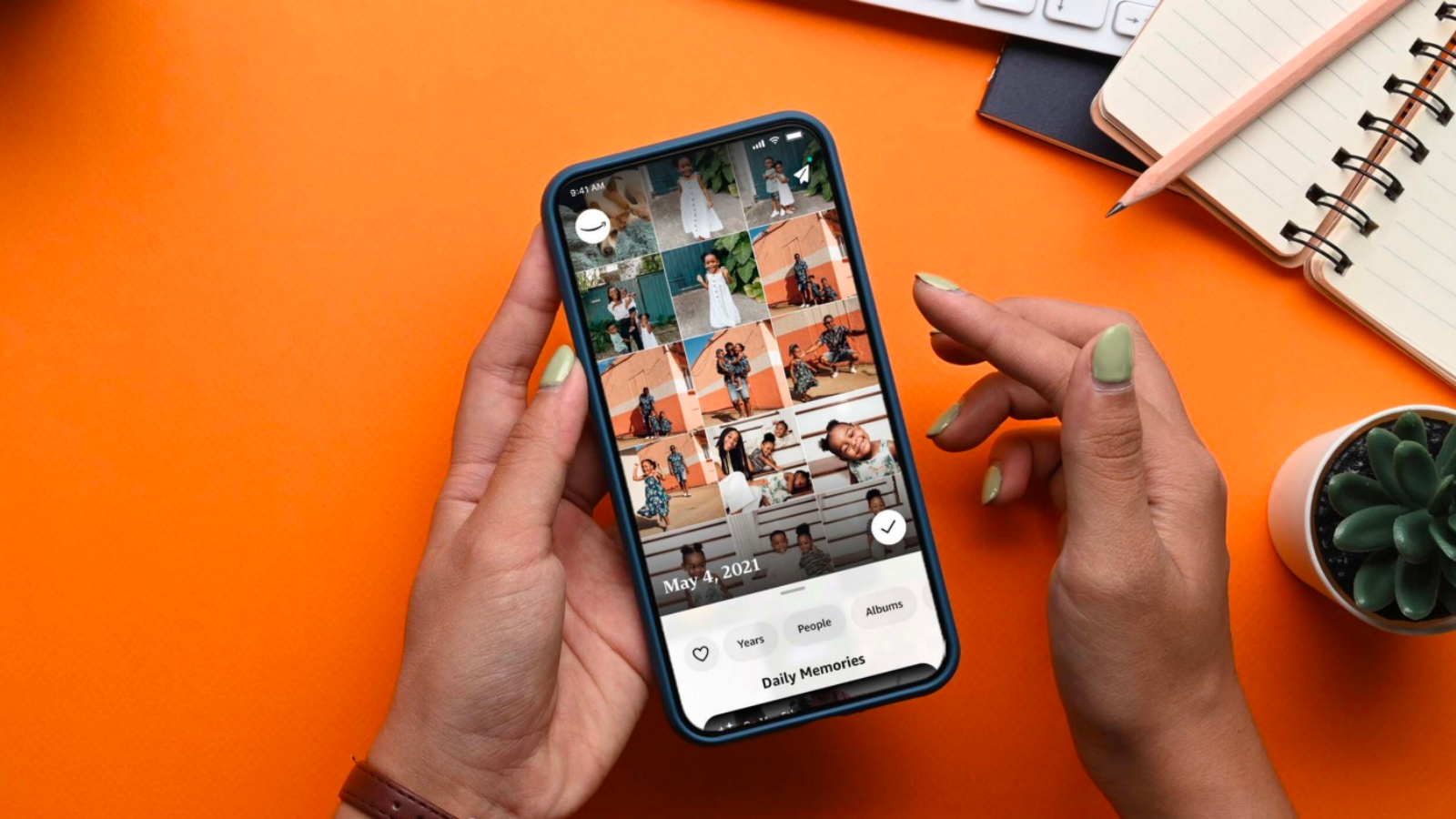 a close-up of a woman with light green nails scrolling through the amazon photos app on an iphone. a cactus and a notebook sit on an orange table next to her hands