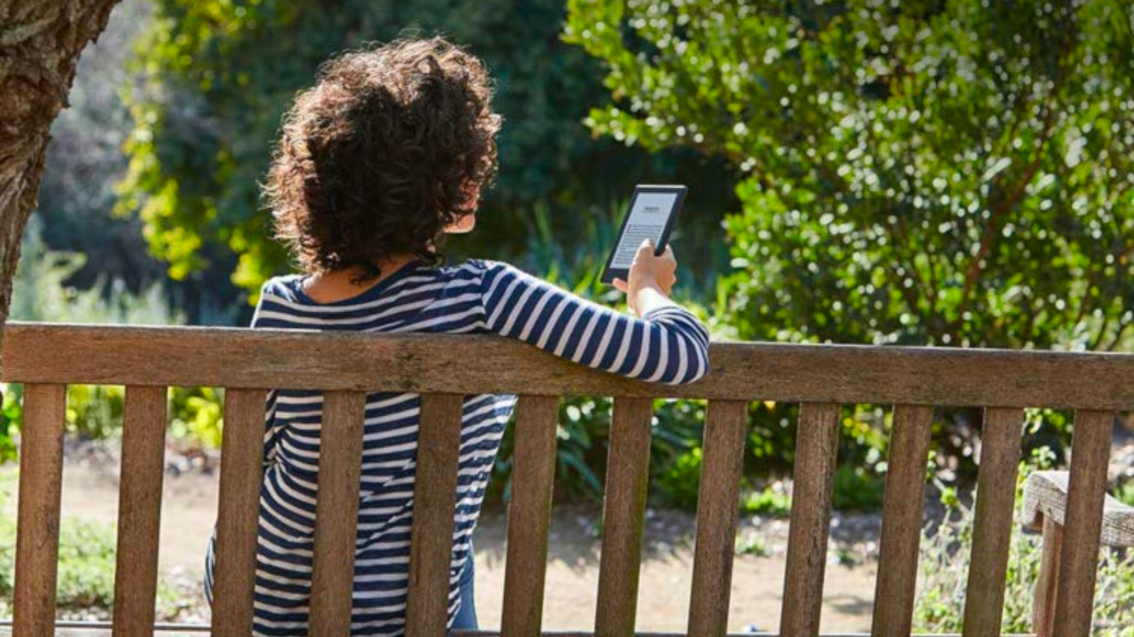 a woman with brown curly hair wearing a striped shirt reads a kindle on a park bench