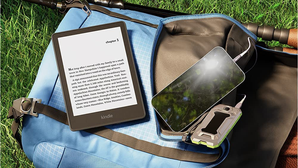 Black Kindle tablet showing half a page of text, The tablet lays next to an iPhone on a blue blanket on top of green grass. 