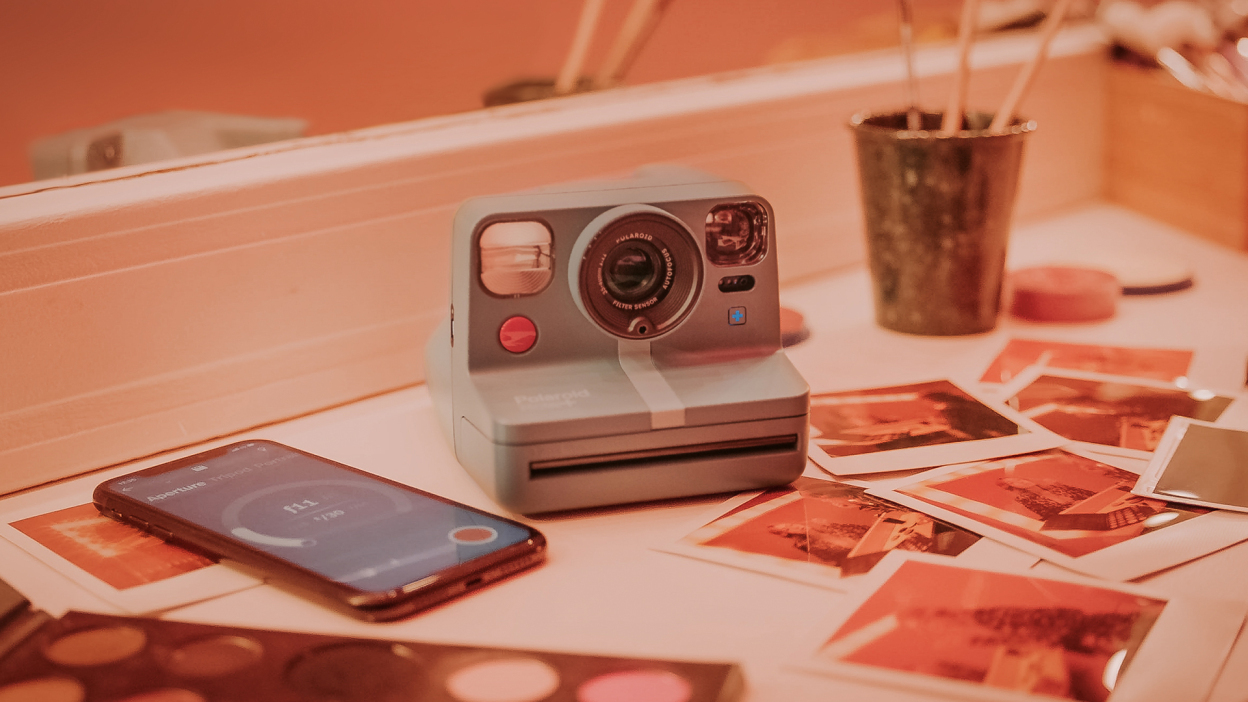 Blue-gray instant camera sits on a table amongst prints and a phone. The image is overlaid with a red tint.