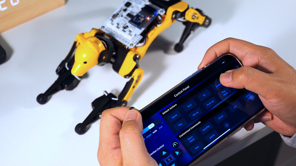 Black and yellow robot dog model on table and hands holding phone displaying control panel