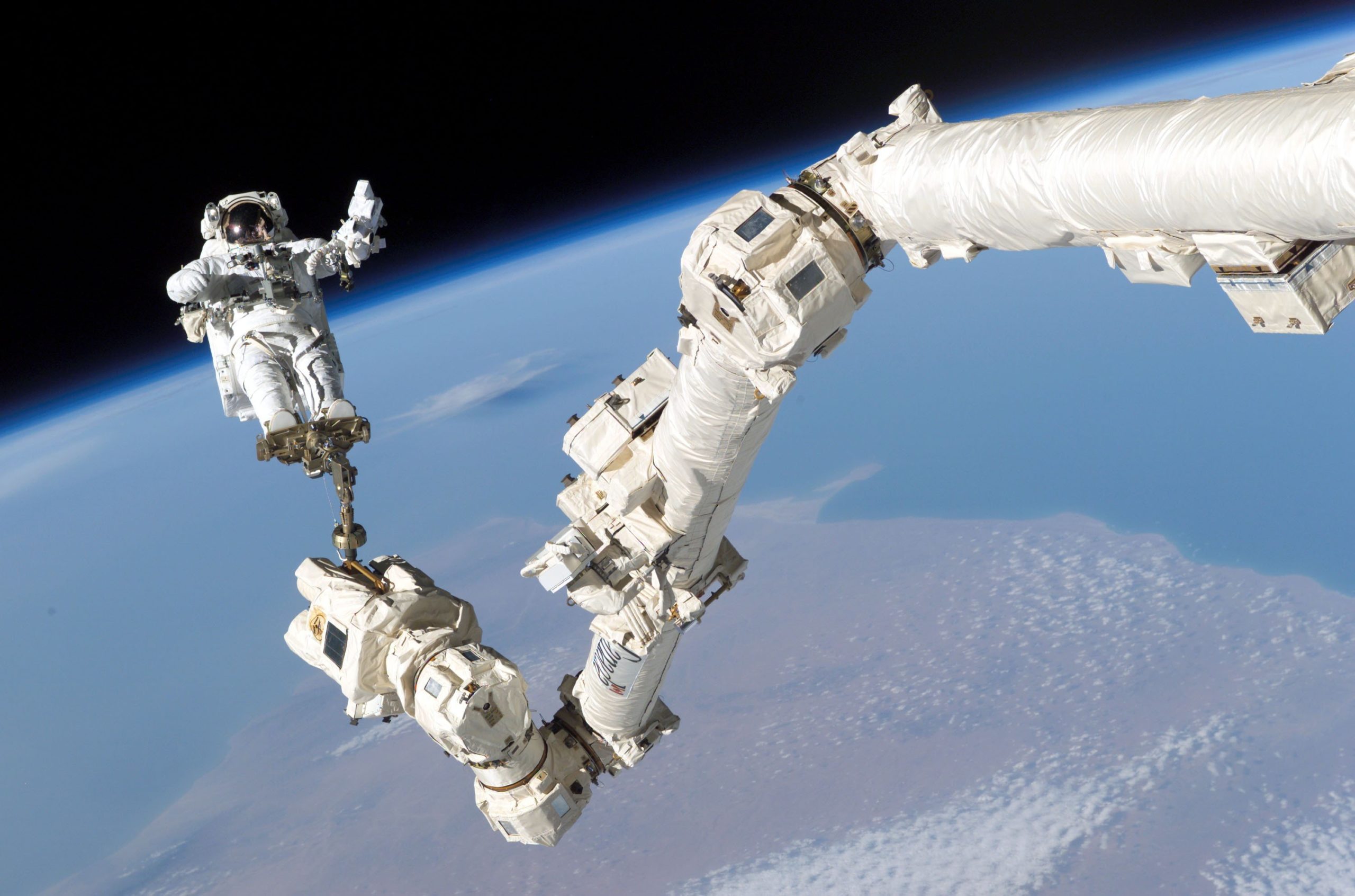 An astronaut floats in space, tethered to the space station, as they complete work outside of it. Behind them you can see the earth!