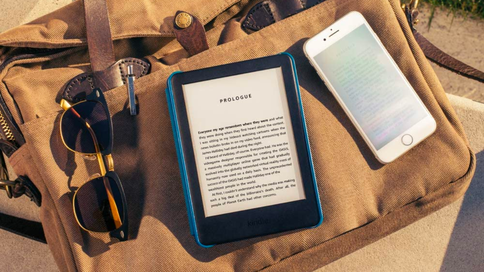 a kindle with a blue cover, an iphone, and a pair of sunglasses resting on a brown canvas bag on a sunny day