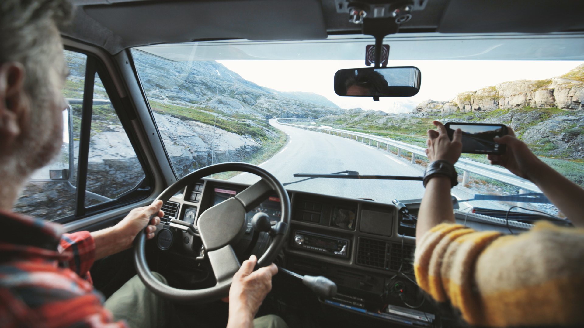 Two people sitting in an RV on a road in the mountains, one driving, one taking a photo on their phone.