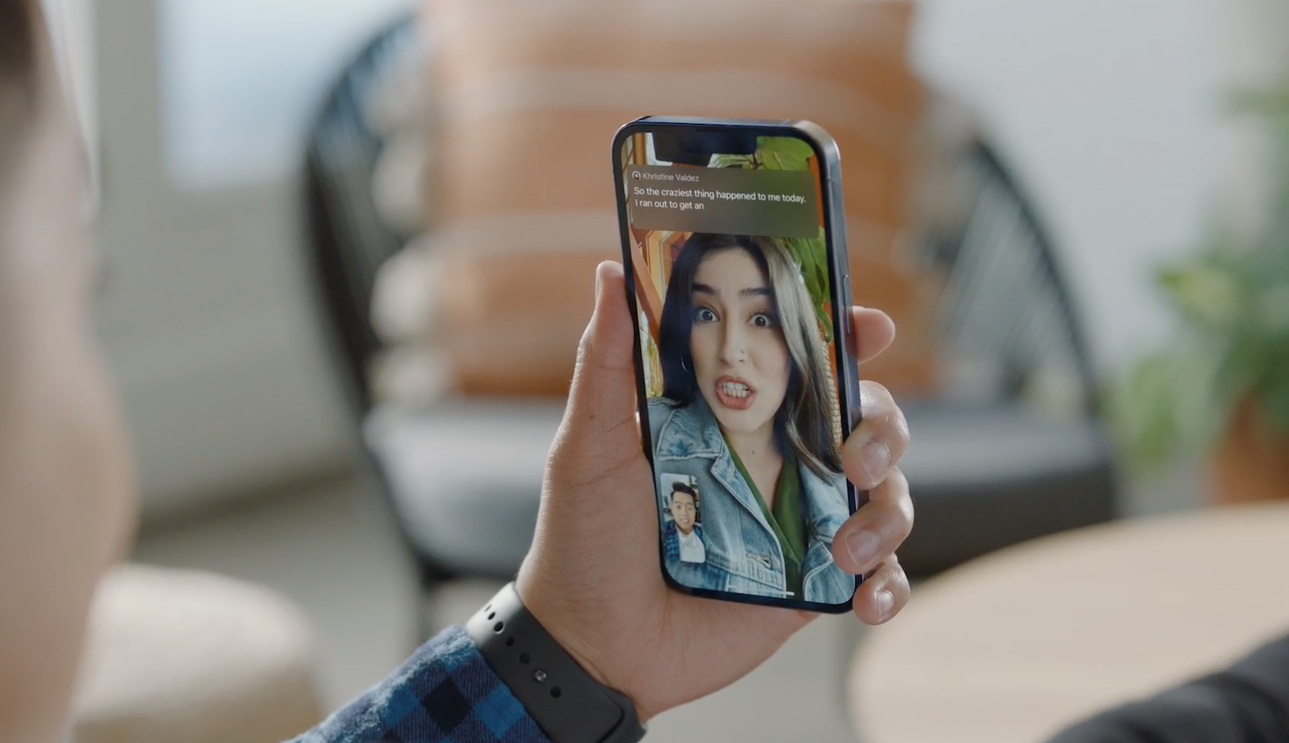 A person holds up an iPhone to their face. The screen shows an active FaceTime call with captions running across the top of the screen above the speaker.