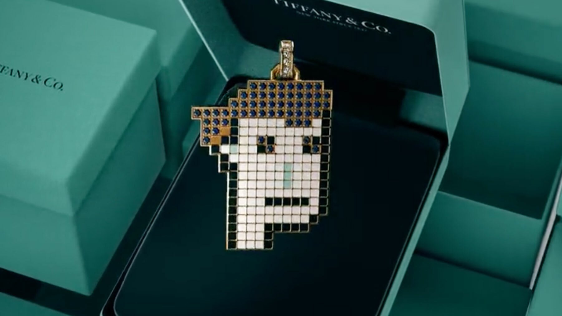 A piece of jewellery in the shape of a pixellated face hangs in front of a green Tiffany box.