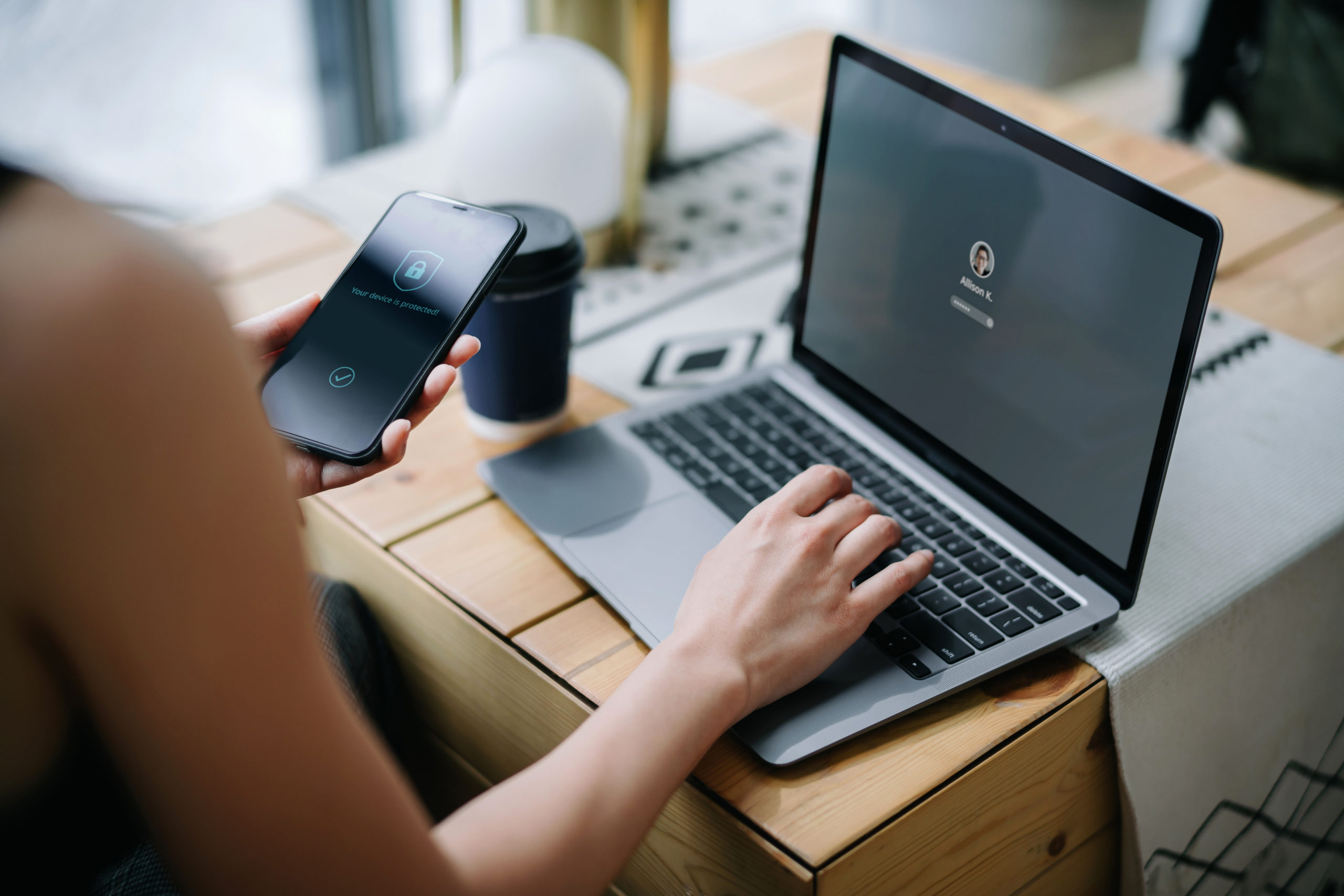 Young businesswoman working on desk, logging in to her laptop and holding smartphone on hand with a security key lock icon on the screen.