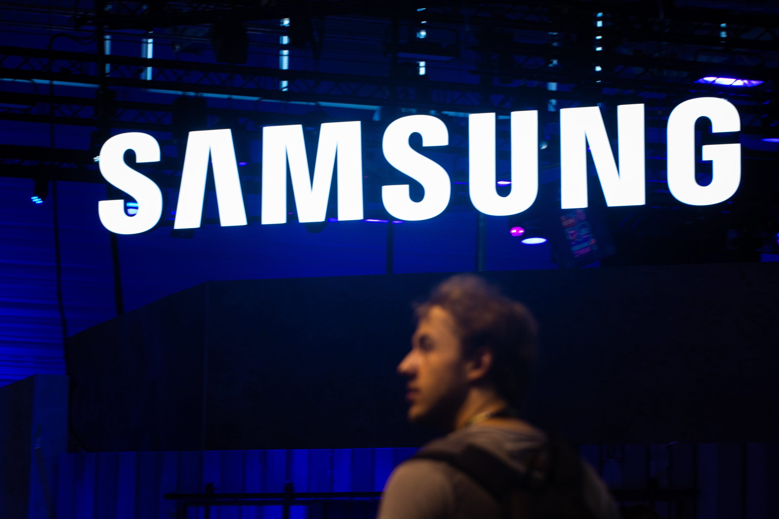 A Samsung sign glowing during the night. A blurry man walks in front of it.