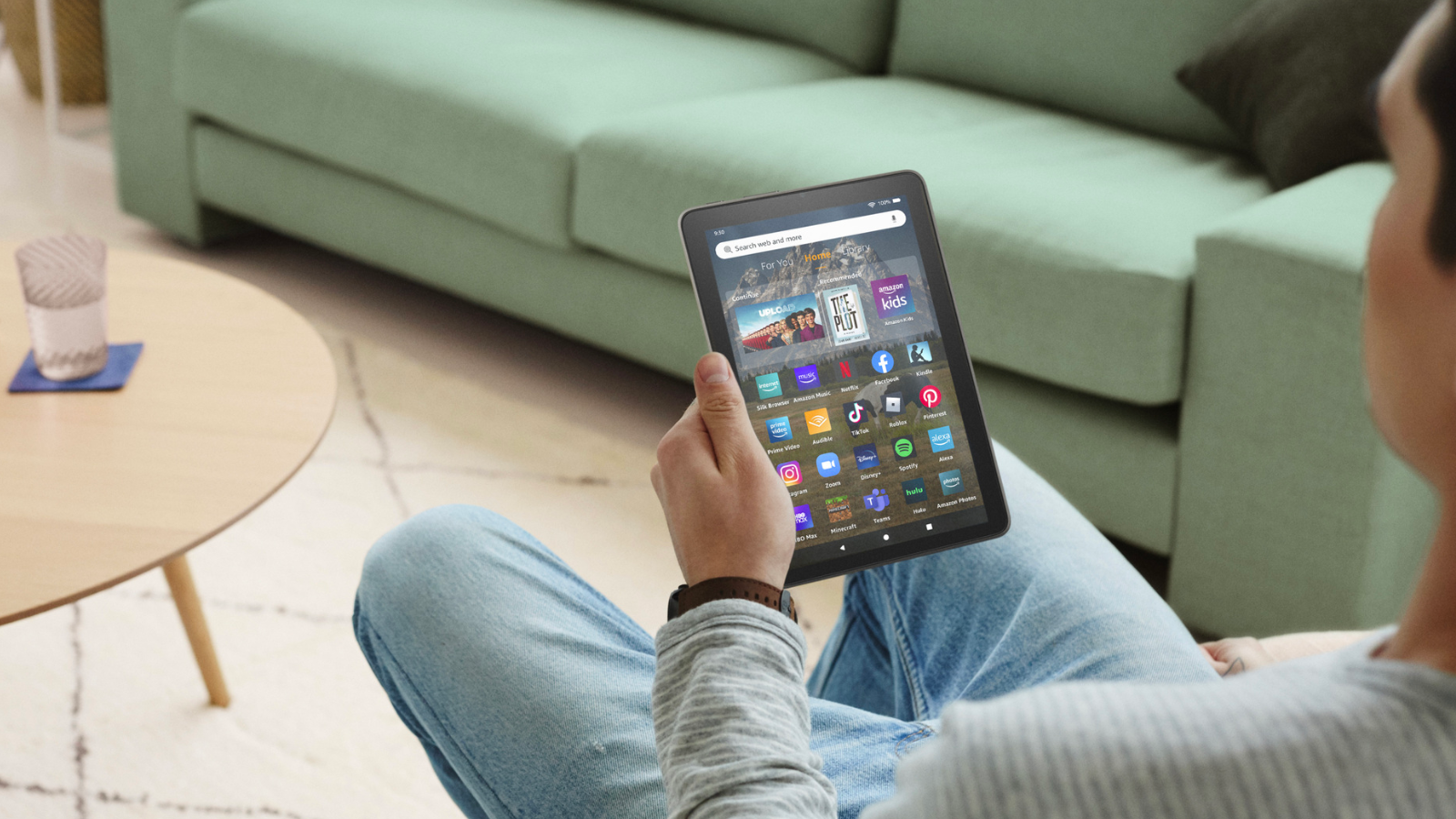 a man in jeans and a gray shirt looks at the dashboard of a fire hd 8 tablet while sitting on a chair in front a wood coffee table and a green couch