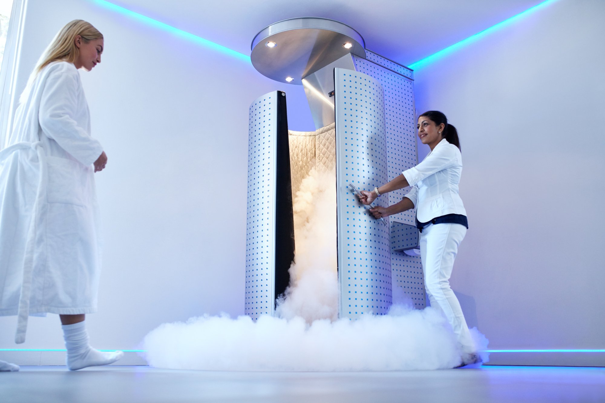 Woman opening a large cryotherapy chamber with smoke billowing out of the bottom. Another woman in a robe approaches.