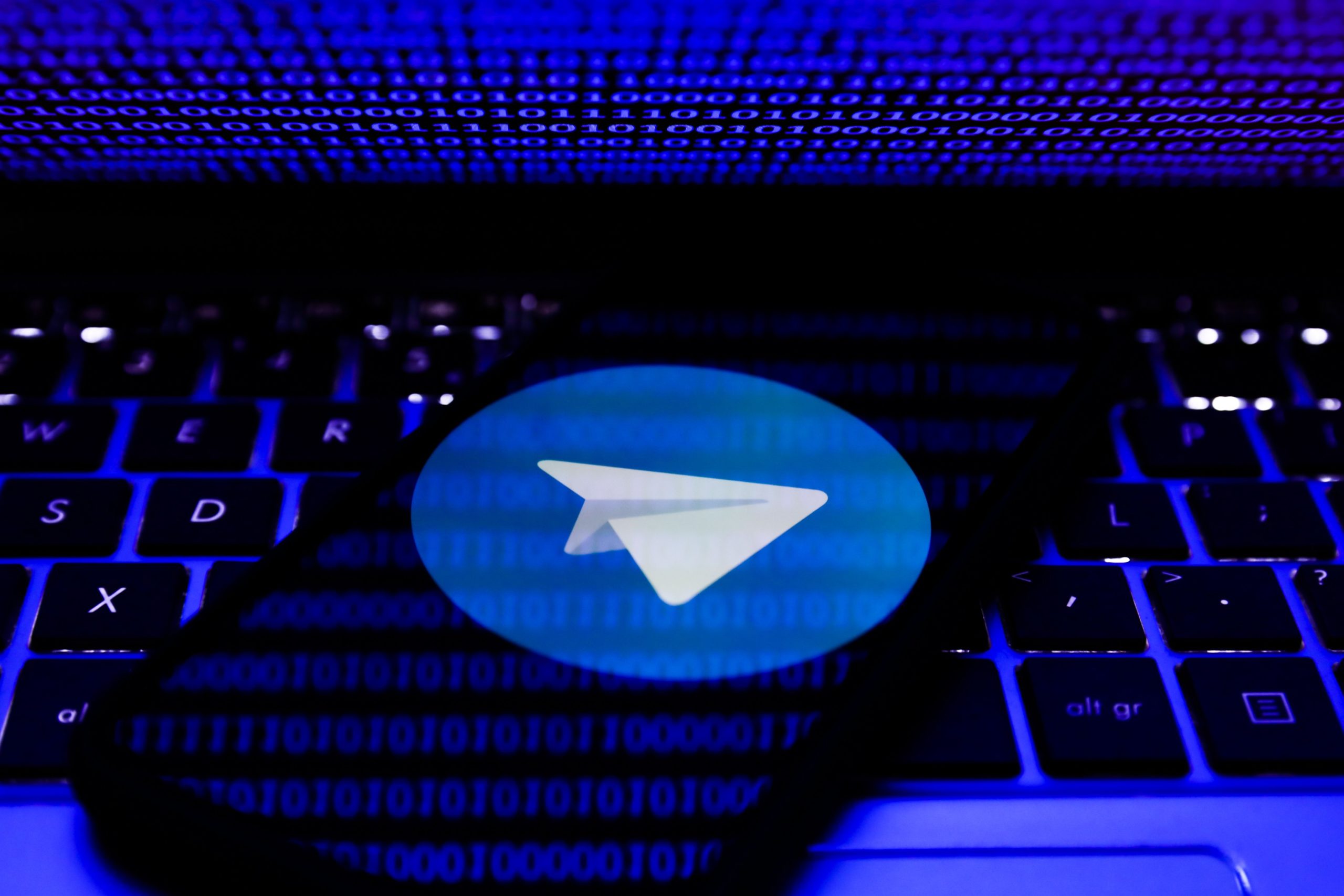 A phone rests on a laptop keyboard. On the phone's screen is the Telegram app's blue and white logo featuring a paper airplane. The reflection of binary code can be seen on the screen, washed in a blue light.
