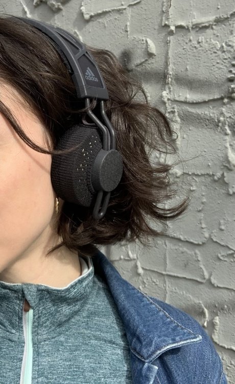A shot of one side of the headphones over the ear of a white woman with short wavy brown hair and small gold hoop earrings, against a blue-grey background.