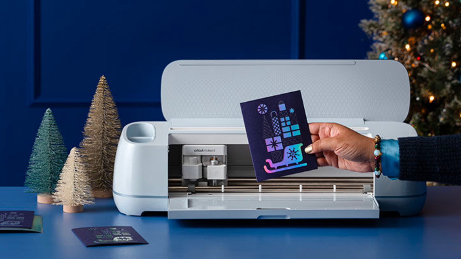  man in a dark blue sweater holding a homemade holiday card in front of a cricut maker 3 machine at a festive holiday table setting