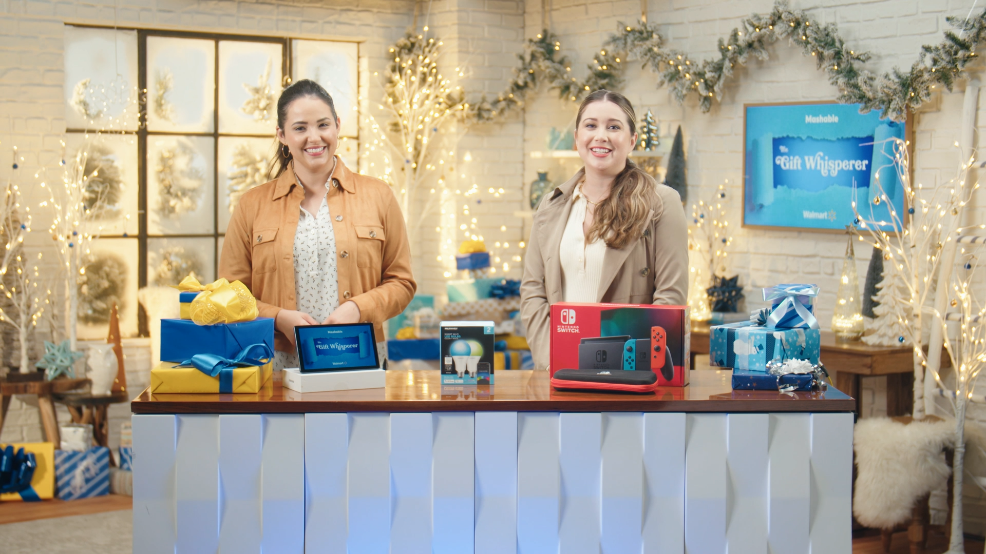 Hosts Cathey Pedrayes and Gaby Ward stand behind table full of holiday gifts from Walmart