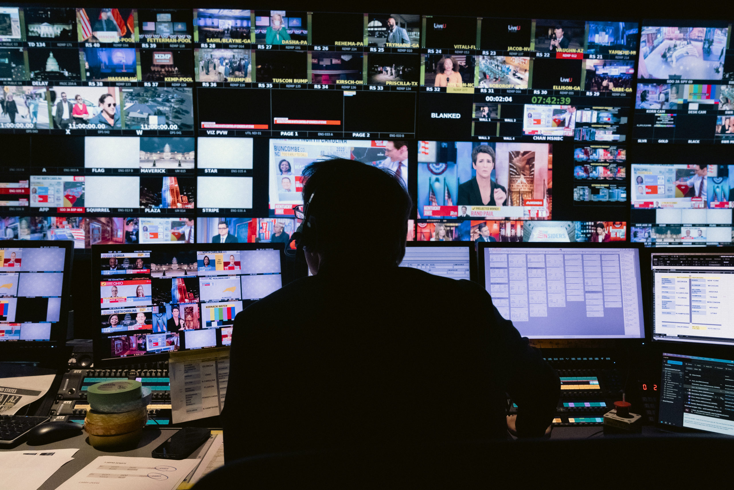 The silhouette of a male from behind. In frotn of him are dozens of small squares with different TV feeds on them.