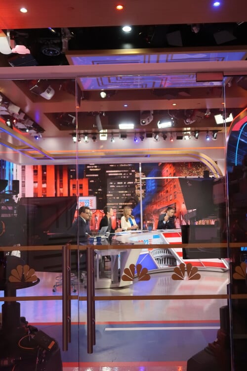 Chris Hayes, Joy Reid, Rachel Maddow, Nicolle Wallace, and Ari Melber at the desk on election night, seen through glass doors and from the side.