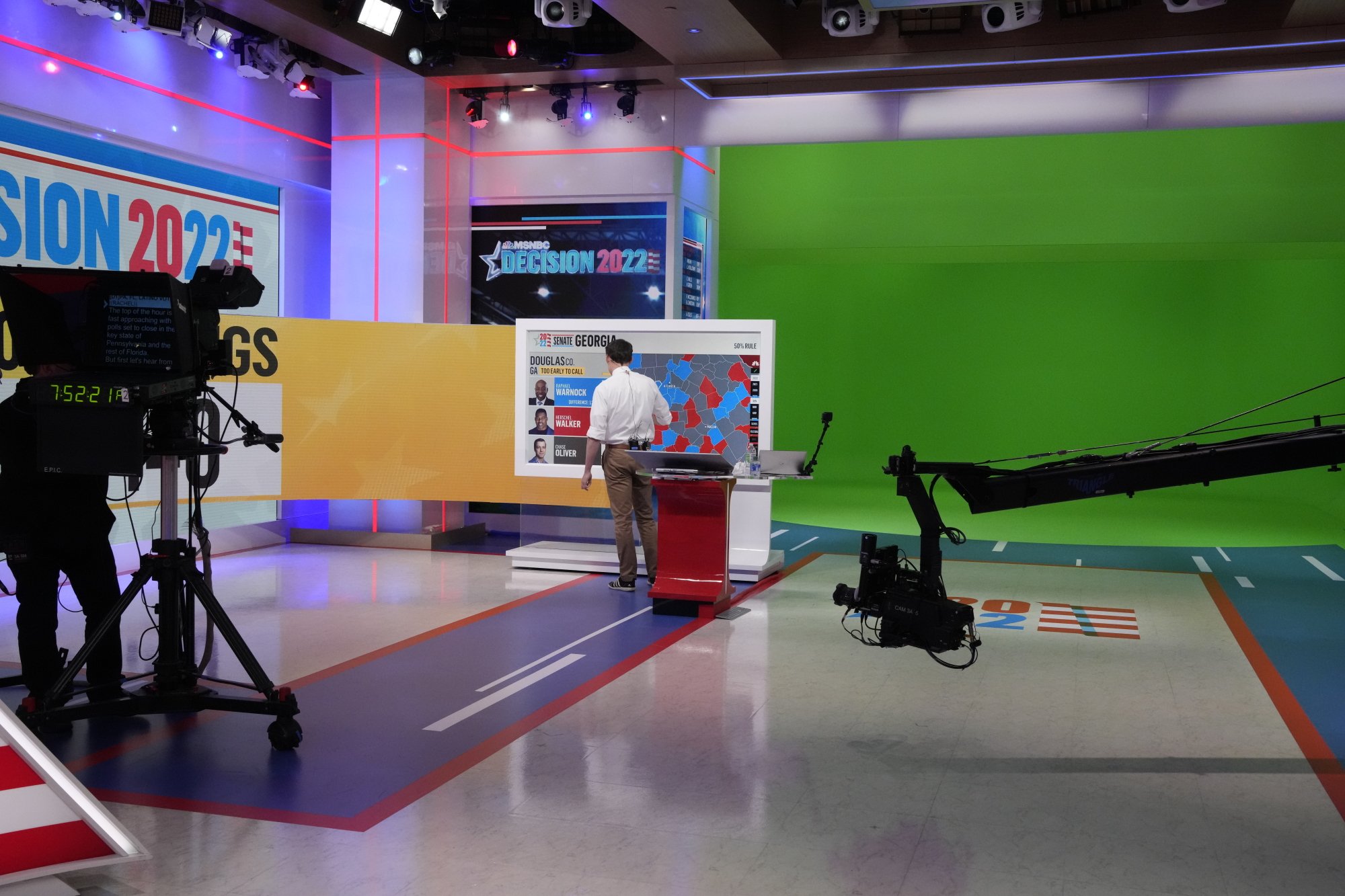 Steve Kornacki stands at his Big Board, filmed by a camera. To the right of him is a large green room.