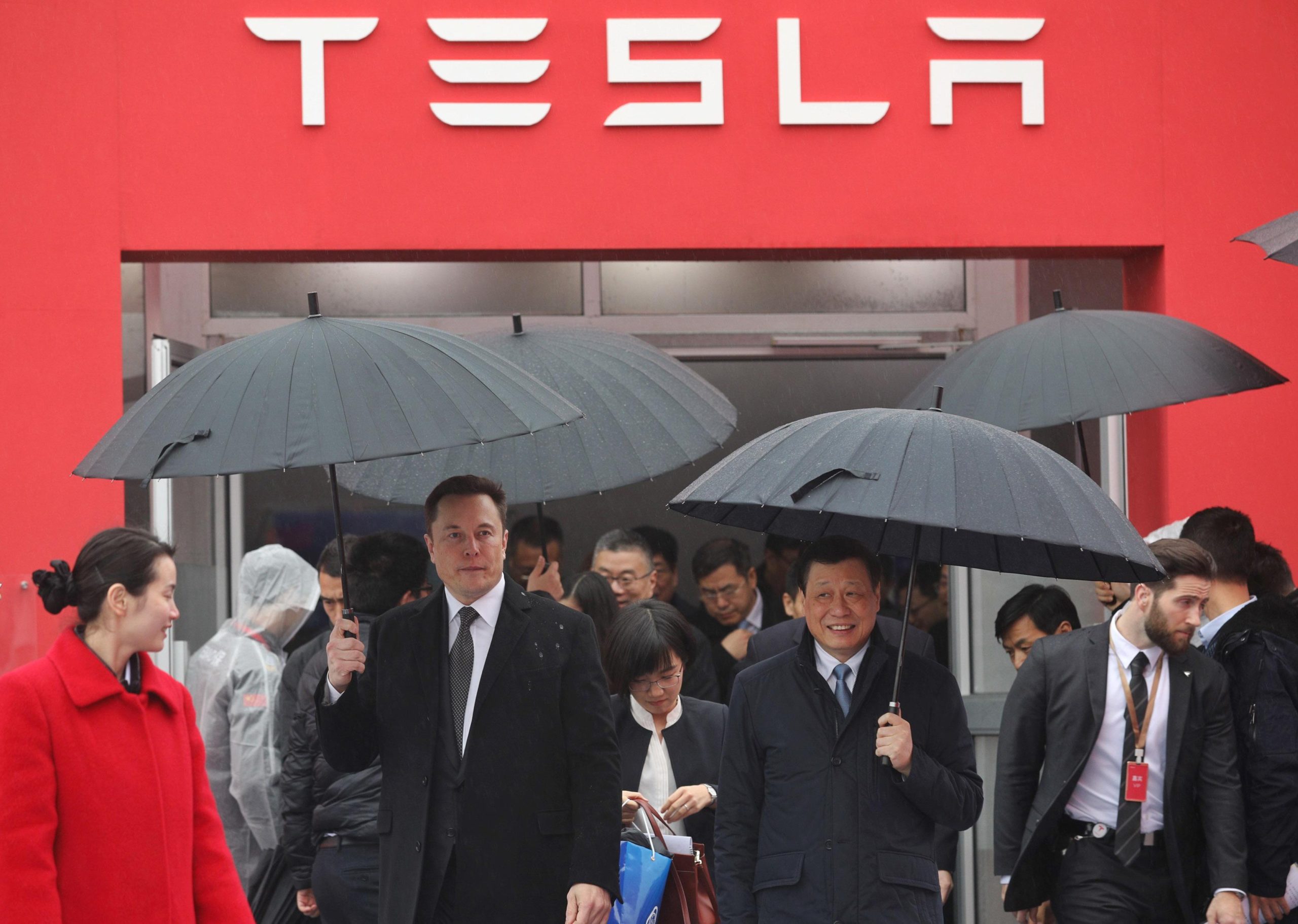 Elon Musk in the rain amid a crowd of people in business attire in front of a Tesla logo on a building