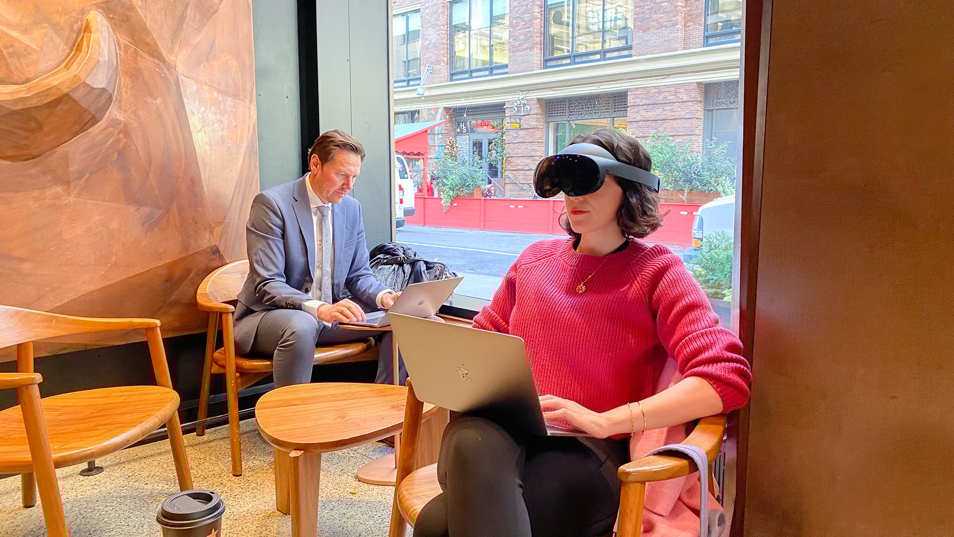 A woman sits in a coffee shop working on a macbook, wearing the meta quest pro. In the background, a man in a business suit works on a laptop.