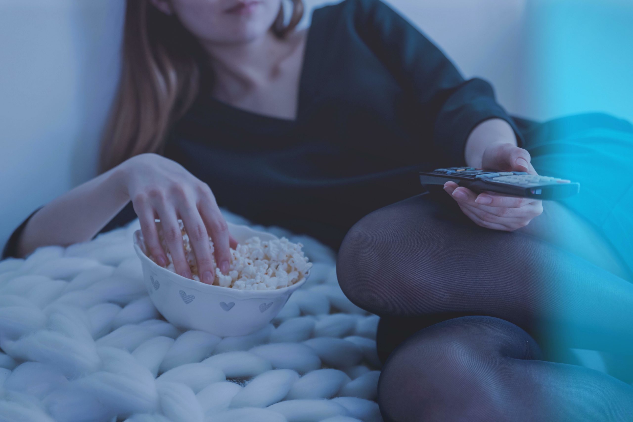 A woman reclines with her right hand in a bowl of popcorn and her left hand holding a remote control.