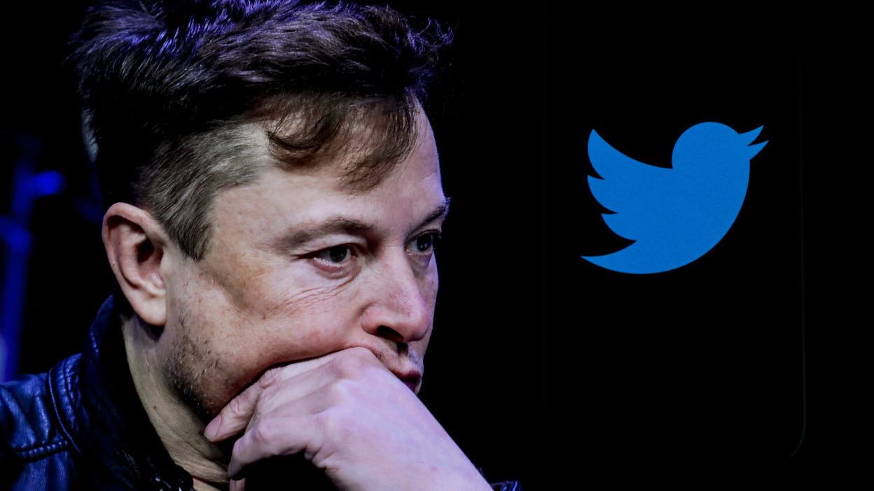 Elon Musk stares concerned with the Twitter logo behind him. 