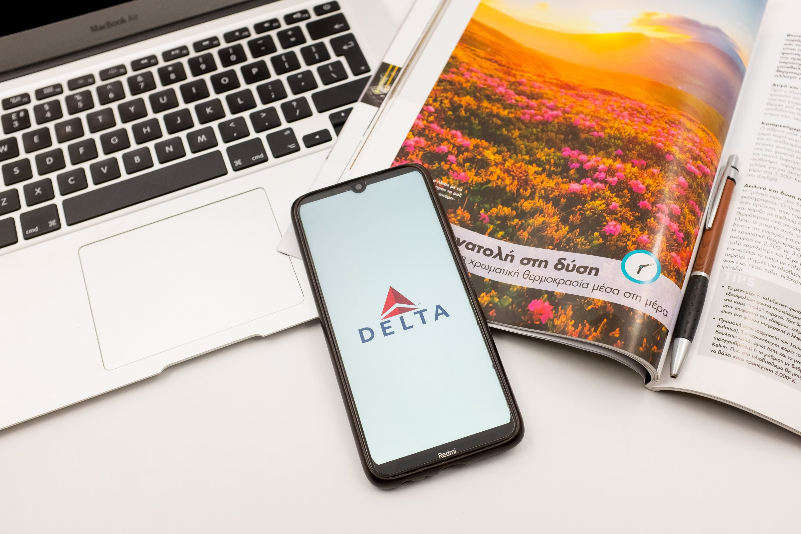 A smartphone sitting on a white surface alongside a laptop and an open magazine.