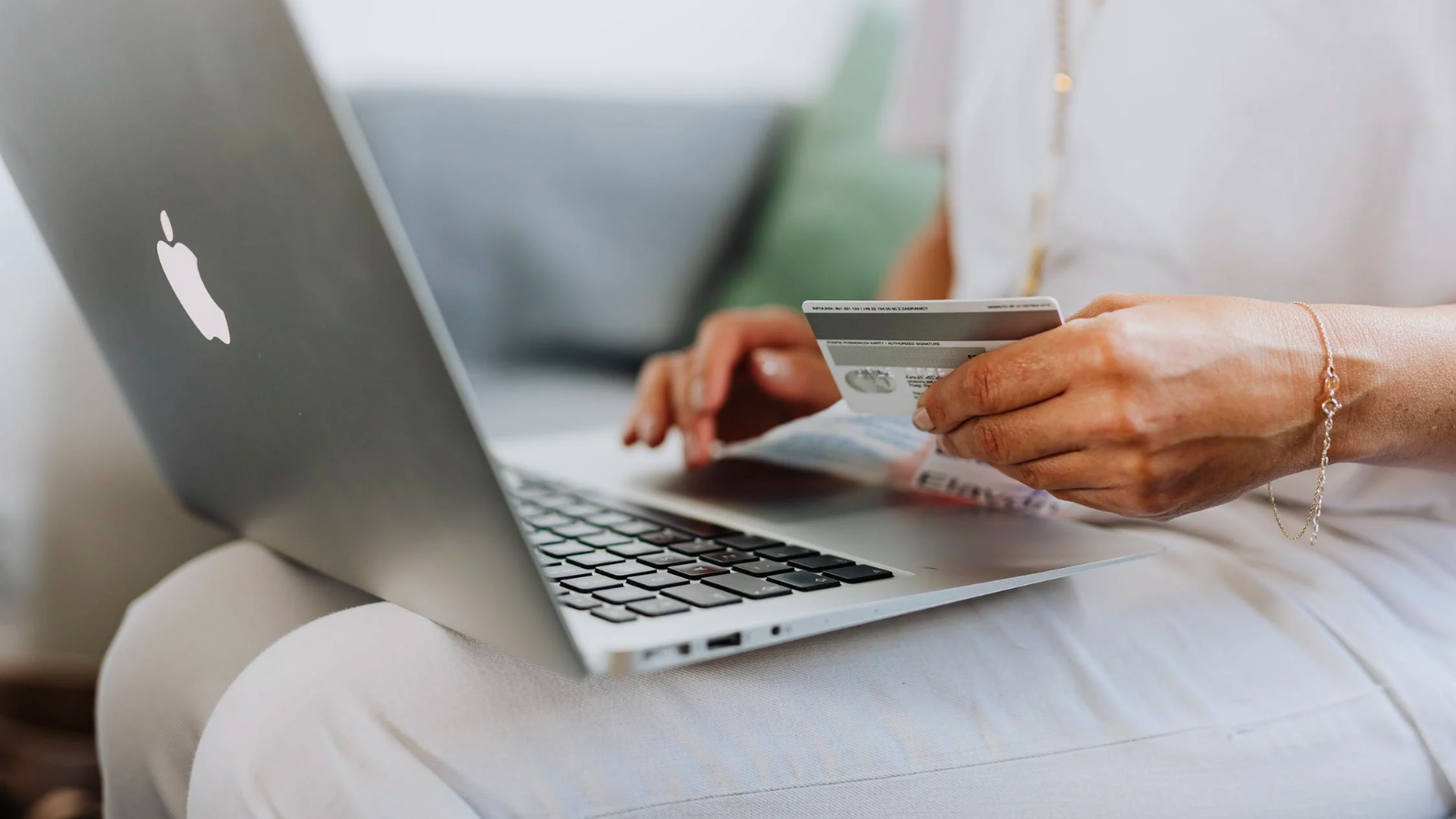 a close-up of a woman in a white top, white pants, and gold jewelry holding a credit card in front of an open macbook